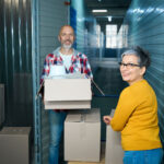 Adult man carries a cardboard box with things while a woman looks at the camera