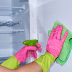 Cleaning empty refrigerator - woman's hands in gloves holding cleaning rag and spray detergent