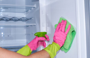 Cleaning empty refrigerator - woman's hands in gloves holding cleaning rag and spray detergent