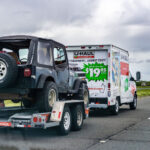 U-Haul truck driving on a road towing a car.