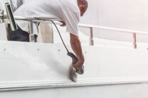 Yacht maintenance. A man polishing side of the white boat by grinder machine in the marina